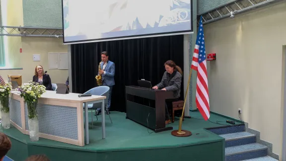 muzyk grający na saksofonie, muzyk grający na pianie, w tle dziewczyna za biurkiem, w tle flaga USA
