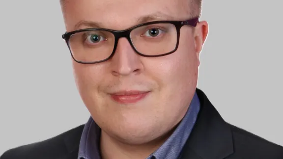 Uśmiechnięty mężczyzna w okularach, ubranym w ciemną marynarkę i błękitną koszulę, pozuje na jednolitym, jasnym tle.