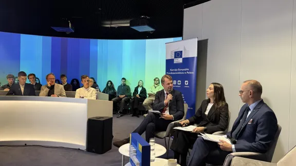 Trzy osoby rozmawiają przy publiczności; obok roll-up Przedstawicielstwa Komisji Europejskiej w Polsce.