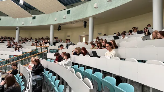 Studenci siedzący w rzędach w nowoczesnej auli wykładowej z jasnymi pulpitami i turkusowymi siedziskami; część osób korzysta z laptopów i tabletów.