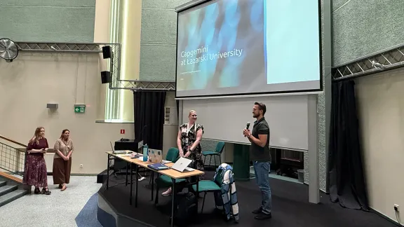 Dwoje prelegentów na scenie auli, mężczyzna mówi do mikrofonu; na ekranie za nimi slajd z napisem „Capgemini at Łazarski University” i logotypem Capgemini. Z boku stoją dwie osoby z obsługi wydarzenia.