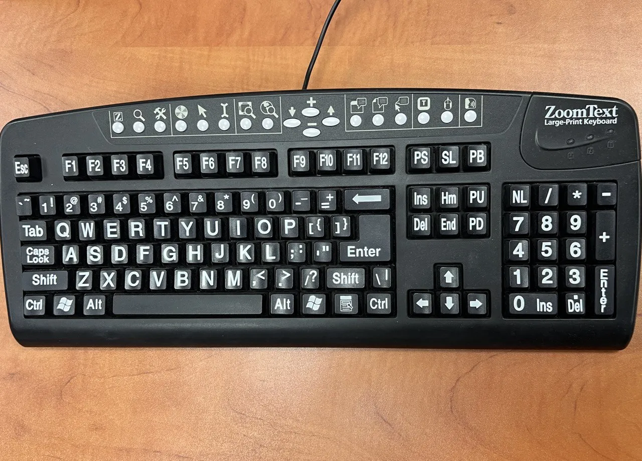 Large black keyboard with white lettering on black enlarged keys. The keyboard has rounded edges. There are 18 additional keys, white „Zoom Text Large-Print Keyboard” inscription and three LEDs at the top.