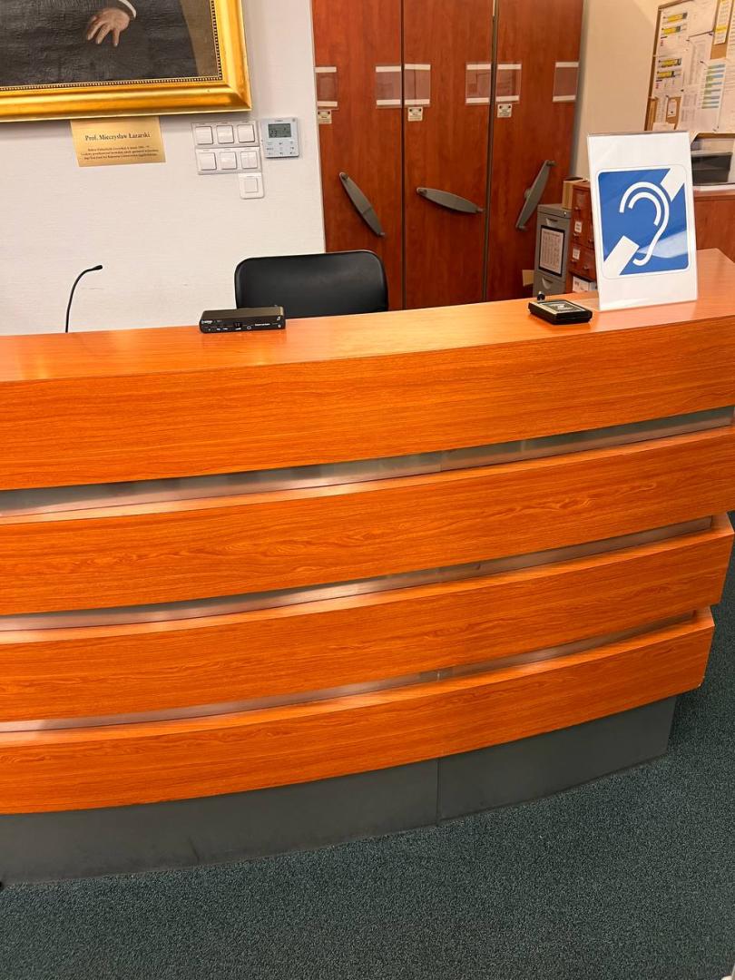 Lada biblioteczna na której umieszczono znak "Pętla indukcyjna". Obok po lewej stronie na ladzie leży elektroniczny czytak legitymacji i wzmacniacz pętli indukcyjnej.