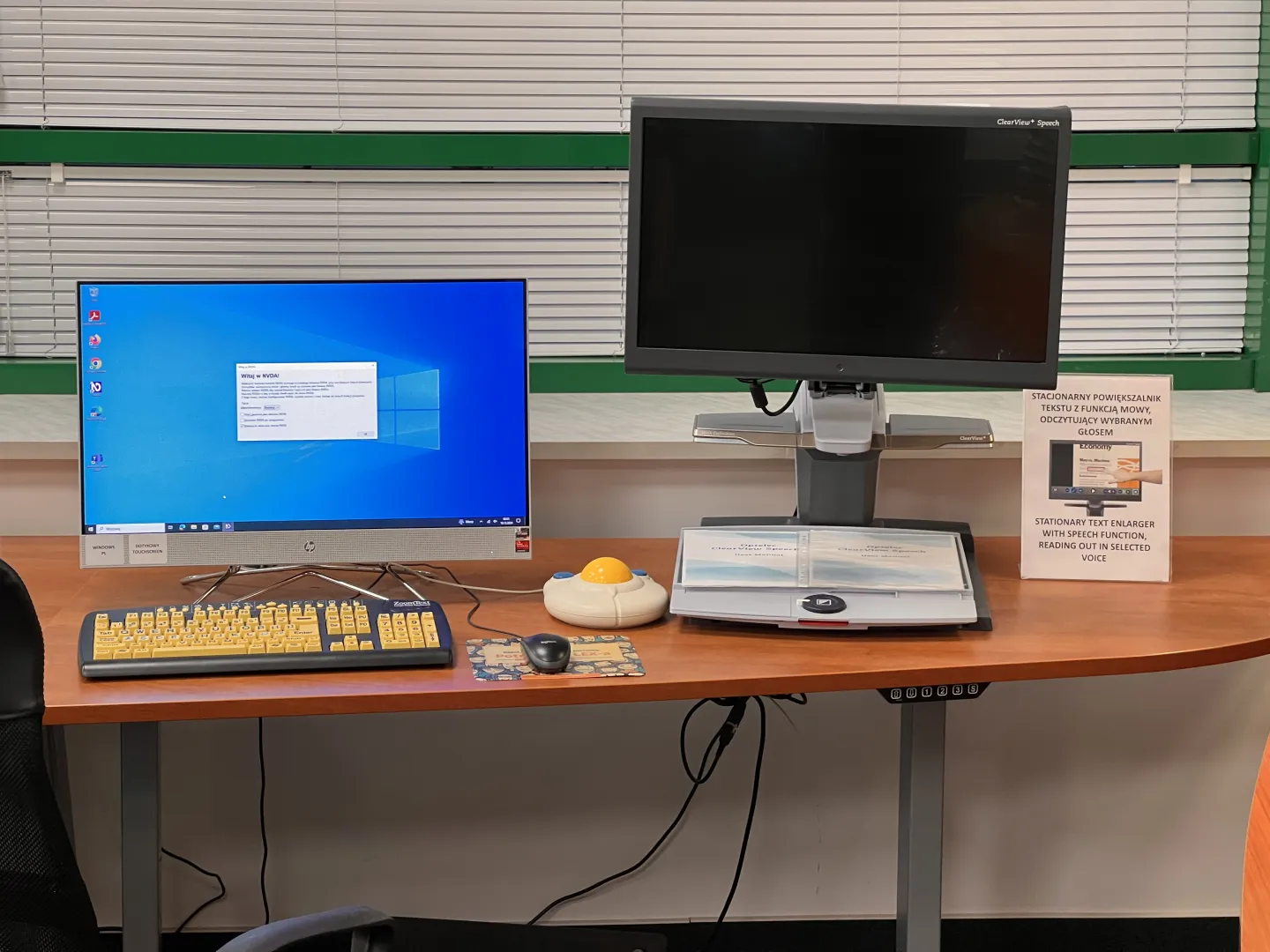 A computer workstation with a touchscreen monitor, a large keyboard with black lettering on yellow keys, and two computer mice - a standard mouse and an enlarged mouse with a yellow trackball and large blue buttons. Under the desk can be seen some cables and a set of buttons that adjust the height of the worktop.