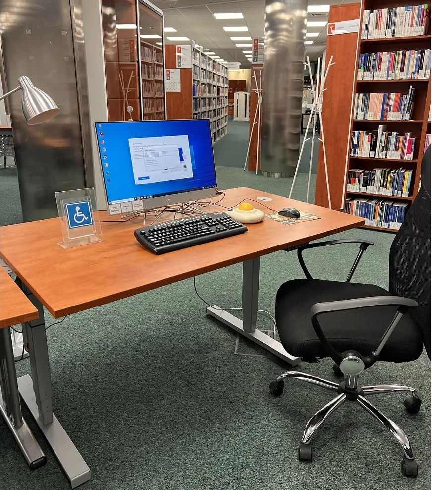 A second computer workstation with a touchscreen monitor, a large keyboard with white lettering on black keys, two computer mice - a standard mouse and an enlarged mouse with a yellow trackball and large blue buttons, and a wheelchair sign on a blue background. Under the desk can be seen two cables and a set of buttons that adjust the height of the work surface. A black computer chair sits next to the desk.