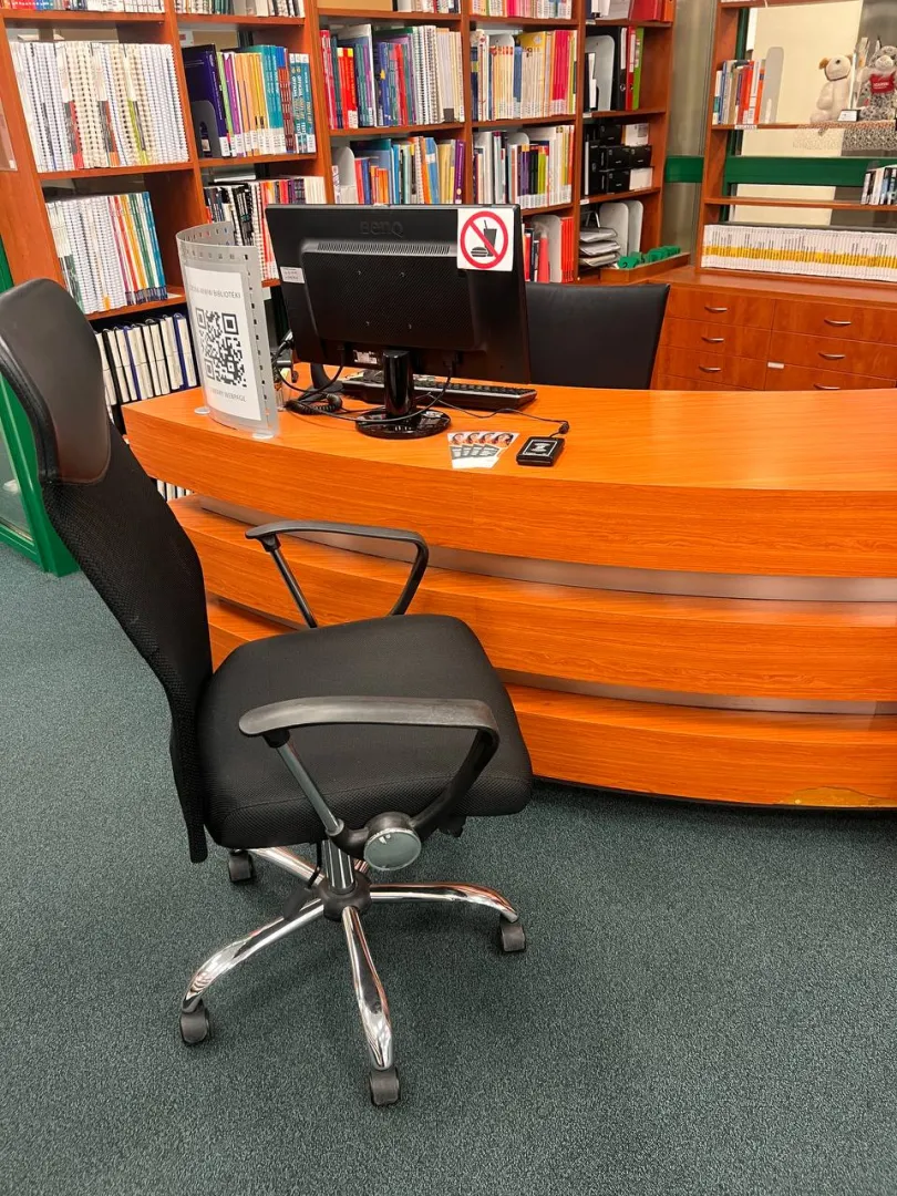Obniżona lada biblioteczna, przed którą jest ustawione krzesło obrotowe. Na ladzie stoi komputer. W tle widać regały z książkami.