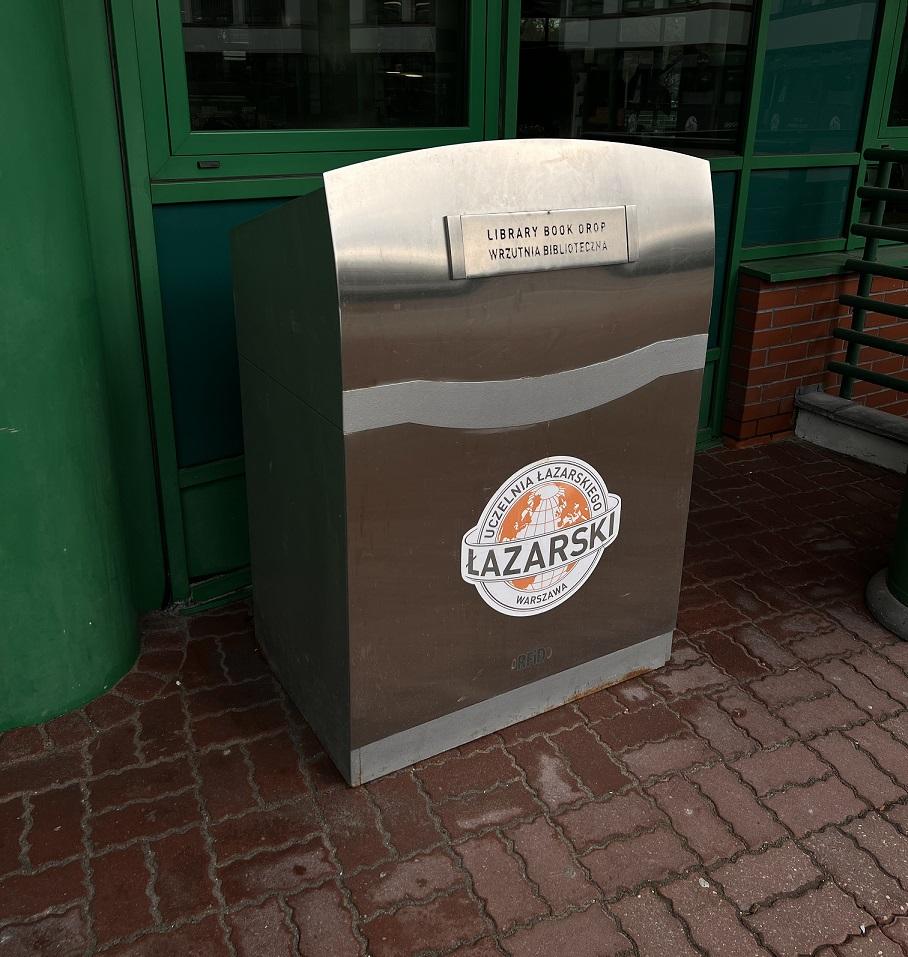 Duży metalowy kontener z logo Uczelni Łazarskiego i napisami "Library Book Drop" oraz "Wrzutnia Biblioteczna".