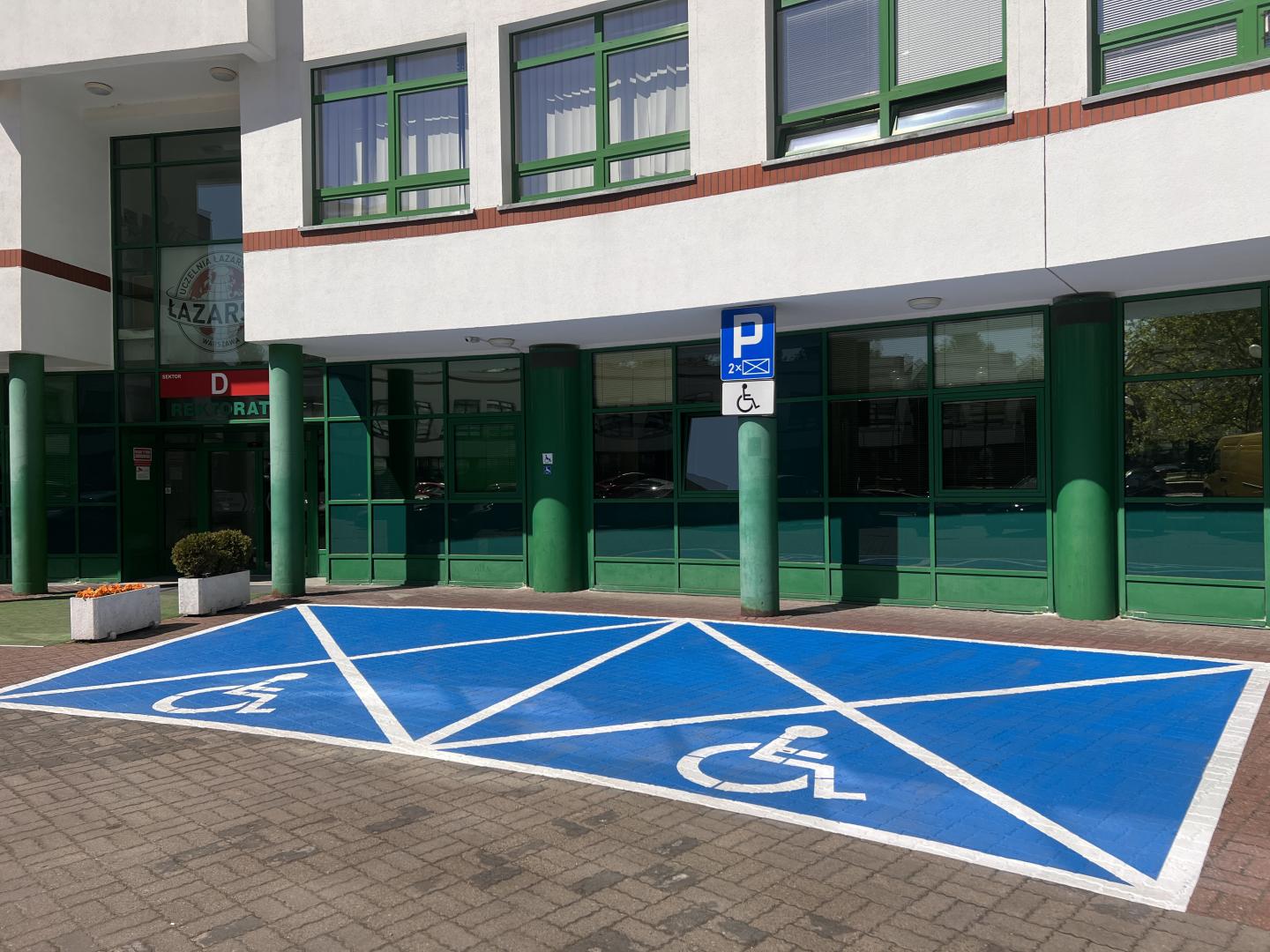 Dwa miejsca parkingowe dla osób z niepełnosprawnościami oznaczone niebieskim kolorem z symbolem wózka inwalidzkiego. W tle widać budynek Uczelni z dużymi oknami oraz wejście oznaczone literą D