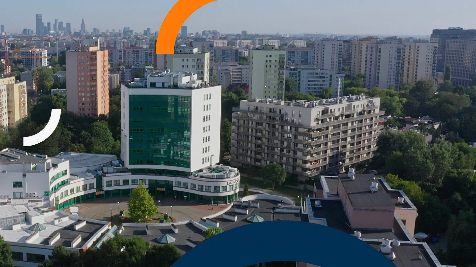 Kampus Akademii Łazarskiego na warszawskim Mokotowie z lotu ptaka: biały budynek z zieloną przeszkloną wieżą otoczony zielenią, w tle panorama Warszawy z Pałacem Kultury i Nauki.