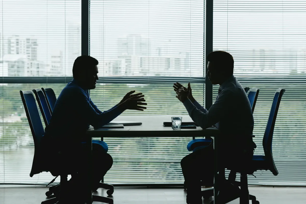 two-human-outlines-against-shuttered-office-window-sitting-opposite-each-other-negotiating