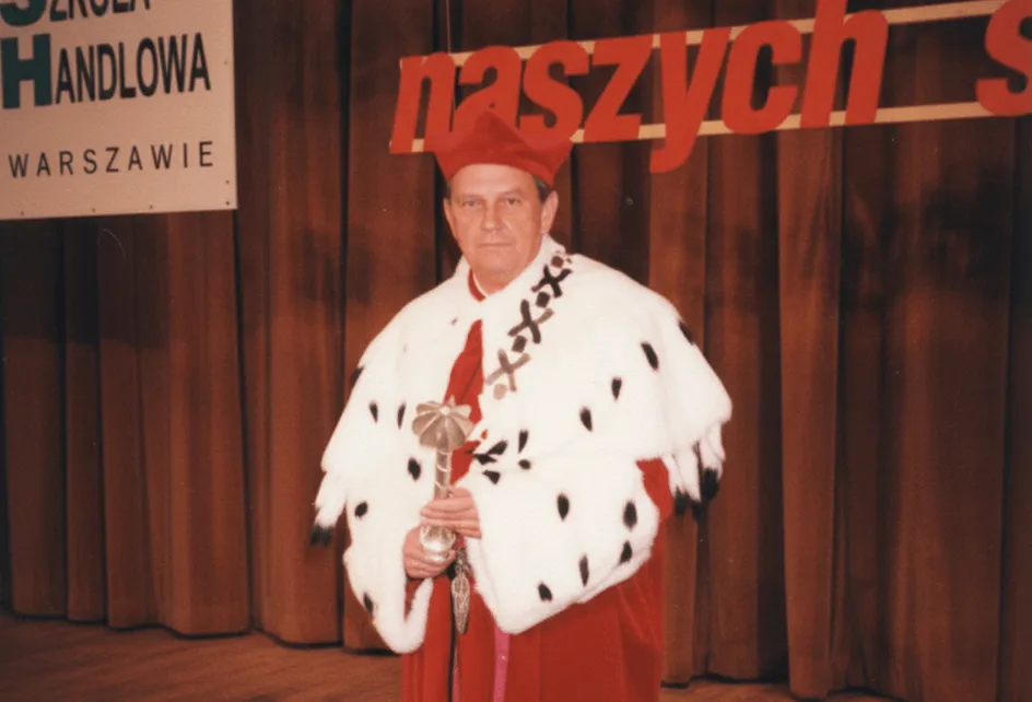Ryszard Łazarski w stroju rektorskim – czerwonej todze, gronostajowej pelerynie i birecie – trzyma berło na tle sceny z napisem „Szkoła Handlowa w Warszawie”.