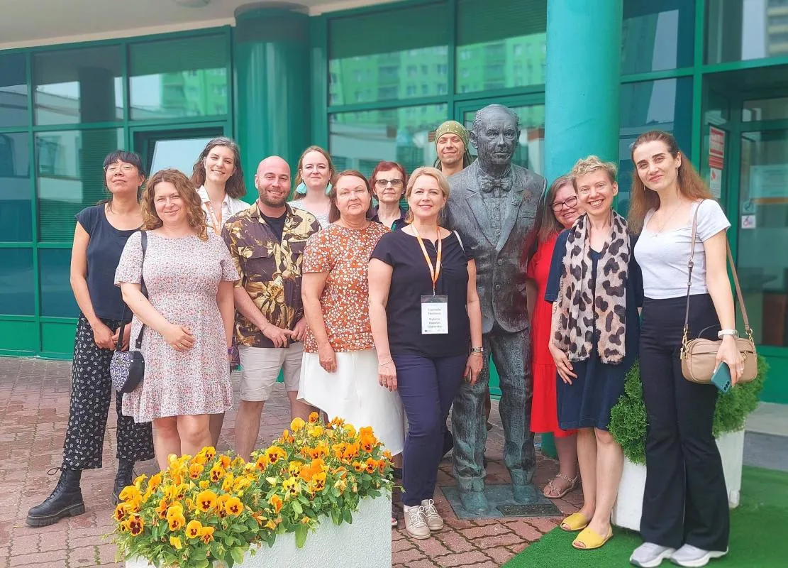 A group of smiling teachers stands in front of the Lazarski University building and poses for a photo at the monument to the founder of the Institution, Ryszard Lazarski.