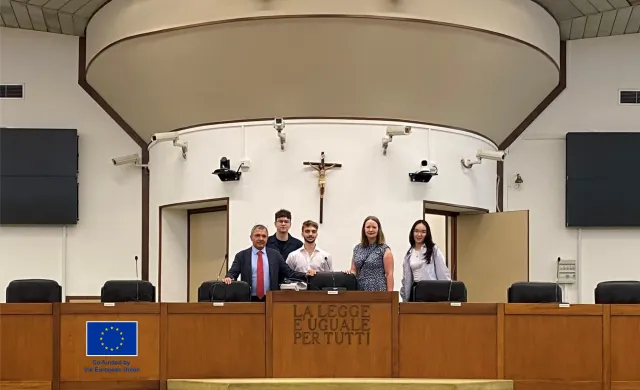 Pięć osób stoi na mównicy i uśmiecha się do kamery. Za nimi na białej ścianie wisi krzyż.