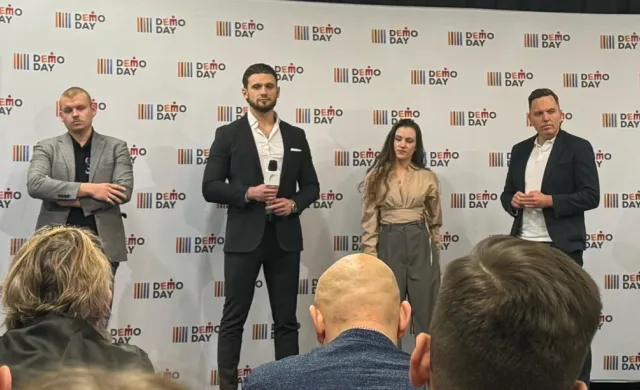 Cztery elegancko ubrane osoby — trzech mężczyzn i jedna kobieta — stoją na tle ścianki z napisem „DEMO DAY”. Na pierwszym planie widoczne są tyły głów kilku osób z publiczności.