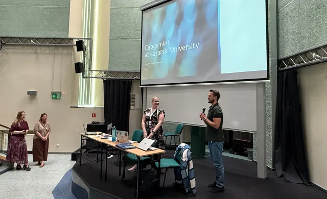 Dwoje prelegentów na scenie auli, mężczyzna mówi do mikrofonu; na ekranie za nimi slajd z napisem „Capgemini at Łazarski University” i logotypem Capgemini. Z boku stoją dwie osoby z obsługi wydarzenia.