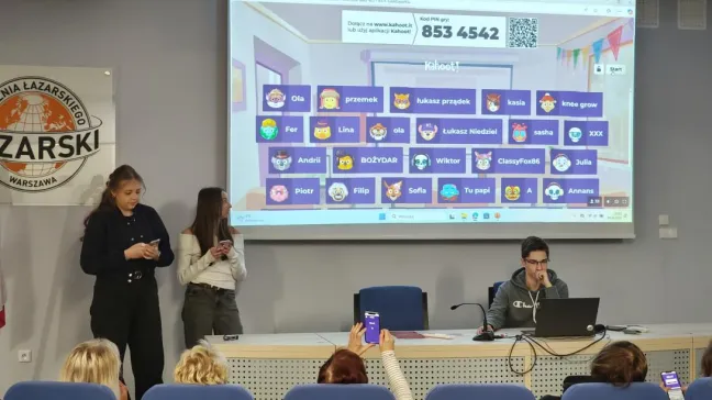 Sala konferencyjna z kilkoma osobami. Na pierwszym planie mężczyzna siedzi przy stole z laptopem, przed nim mikrofon. Na ścianie za nim wyświetlany jest ekran z aplikacją, na którym widoczne są różne awatary i nazwy użytkowników. 853 4542. W tle, na podwyższeniu, stoją dwie kobiety z telefonami w rękach. W sali znajdują się rzędy niebieskich foteli, na których siedzą inne osoby.