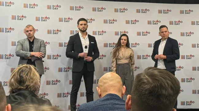 Cztery elegancko ubrane osoby — trzech mężczyzn i jedna kobieta — stoją na tle ścianki z napisem „DEMO DAY”. Na pierwszym planie widoczne są tyły głów kilku osób z publiczności.