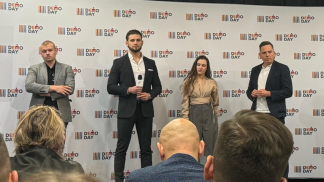 Cztery elegancko ubrane osoby — trzech mężczyzn i jedna kobieta — stoją na tle ścianki z napisem „DEMO DAY”. Na pierwszym planie widoczne są tyły głów kilku osób z publiczności.