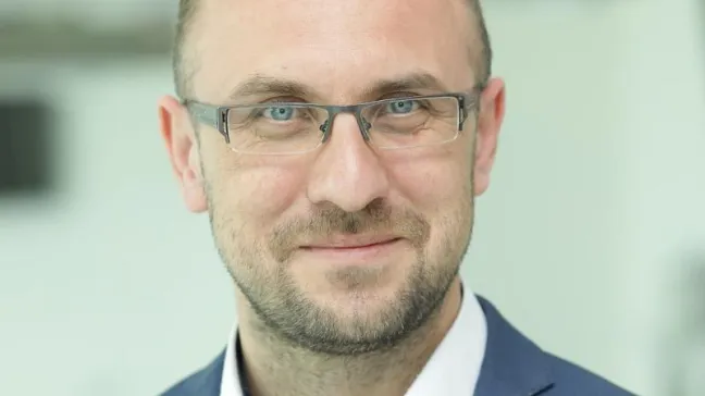 Zdjęcie przedstawia mężczyznę w średnim wieku, który nosi okulary. Ma krótkie, ciemne włosy i lekki zarost. Ubrany jest w niebieską marynarkę i białą koszulę. Mężczyzna patrzy prosto w obiektyw i lekko się uśmiecha. Tło jest rozmyte i jasne, co sprawia, że postać jest dobrze widoczna.