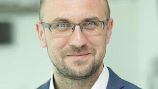 Zdjęcie przedstawia mężczyznę w średnim wieku, który nosi okulary. Ma krótkie, ciemne włosy i lekki zarost. Ubrany jest w niebieską marynarkę i białą koszulę. Mężczyzna patrzy prosto w obiektyw i lekko się uśmiecha. Tło jest rozmyte i jasne, co sprawia, że postać jest dobrze widoczna.