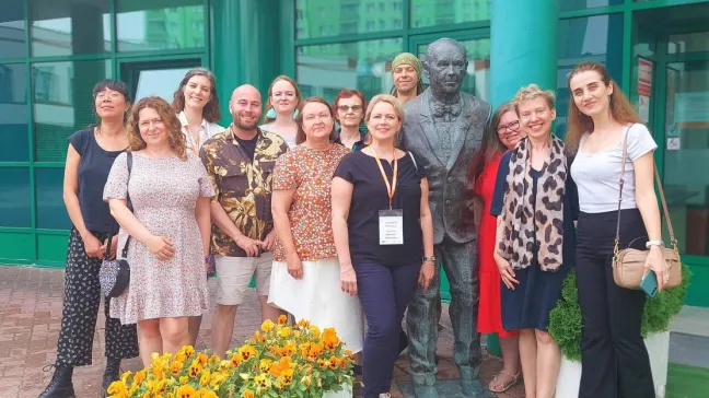 A group of smiling teachers stands in front of the Lazarski University building and poses for a photo at the monument to the founder of the Institution, Ryszard Lazarski.
