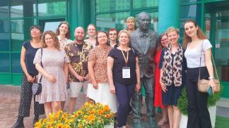 A group of smiling teachers stands in front of the Lazarski University building and poses for a photo at the monument to the founder of the Institution, Ryszard Lazarski.