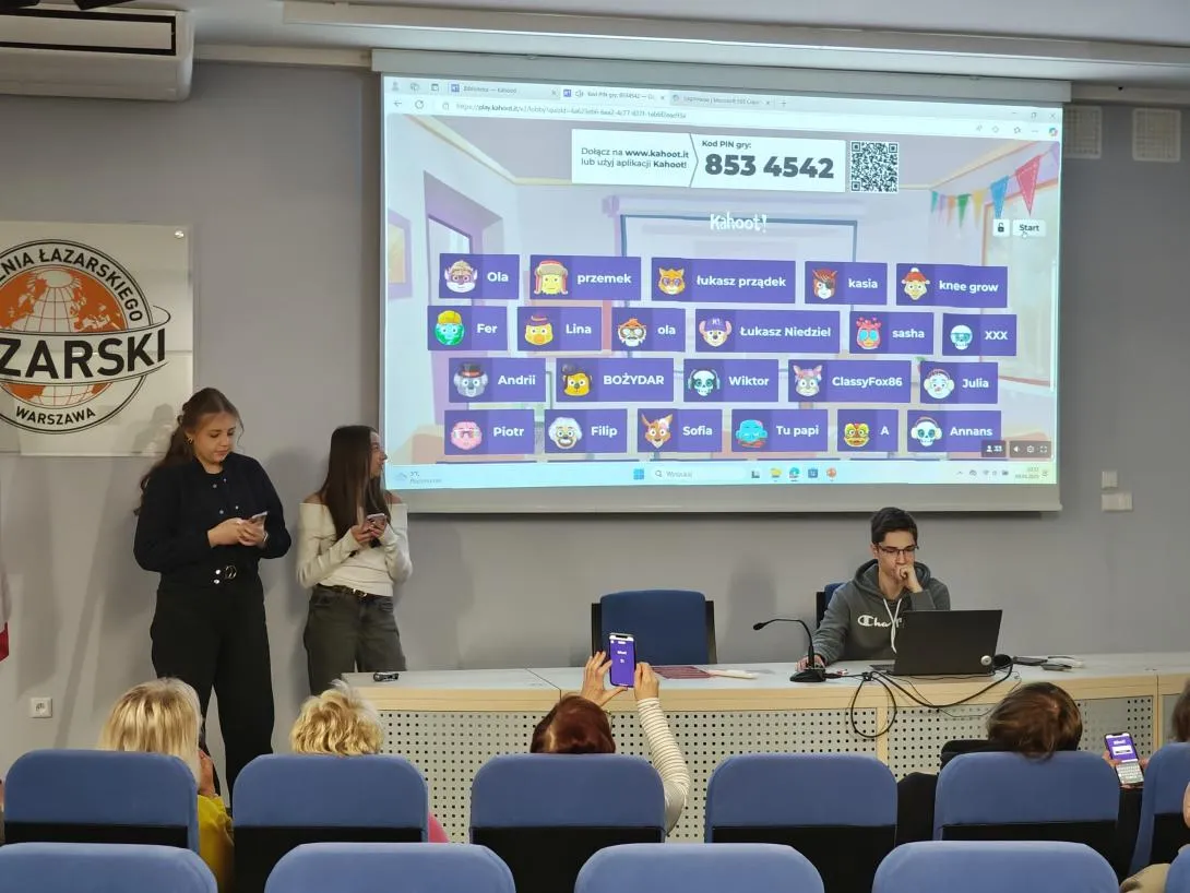 Sala konferencyjna z kilkoma osobami. Na pierwszym planie mężczyzna siedzi przy stole z laptopem, przed nim mikrofon. Na ścianie za nim wyświetlany jest ekran z aplikacją, na którym widoczne są różne awatary i nazwy użytkowników. 853 4542. W tle, na podwyższeniu, stoją dwie kobiety z telefonami w rękach. W sali znajdują się rzędy niebieskich foteli, na których siedzą inne osoby.