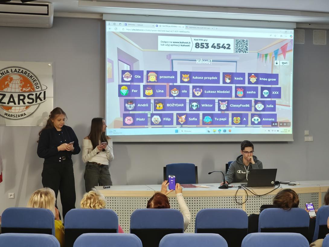 Sala konferencyjna z kilkoma osobami. Na pierwszym planie mężczyzna siedzi przy stole z laptopem, przed nim mikrofon. Na ścianie za nim wyświetlany jest ekran z aplikacją, na którym widoczne są różne awatary i nazwy użytkowników. 853 4542. W tle, na podwyższeniu, stoją dwie kobiety z telefonami w rękach. W sali znajdują się rzędy niebieskich foteli, na których siedzą inne osoby.