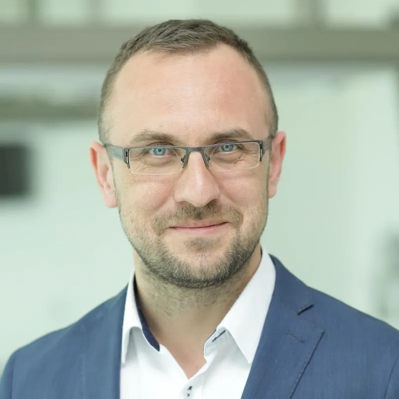 Zdjęcie przedstawia mężczyznę w średnim wieku, który nosi okulary. Ma krótkie, ciemne włosy i lekki zarost. Ubrany jest w niebieską marynarkę i białą koszulę. Mężczyzna patrzy prosto w obiektyw i lekko się uśmiecha. Tło jest rozmyte i jasne, co sprawia, że postać jest dobrze widoczna.