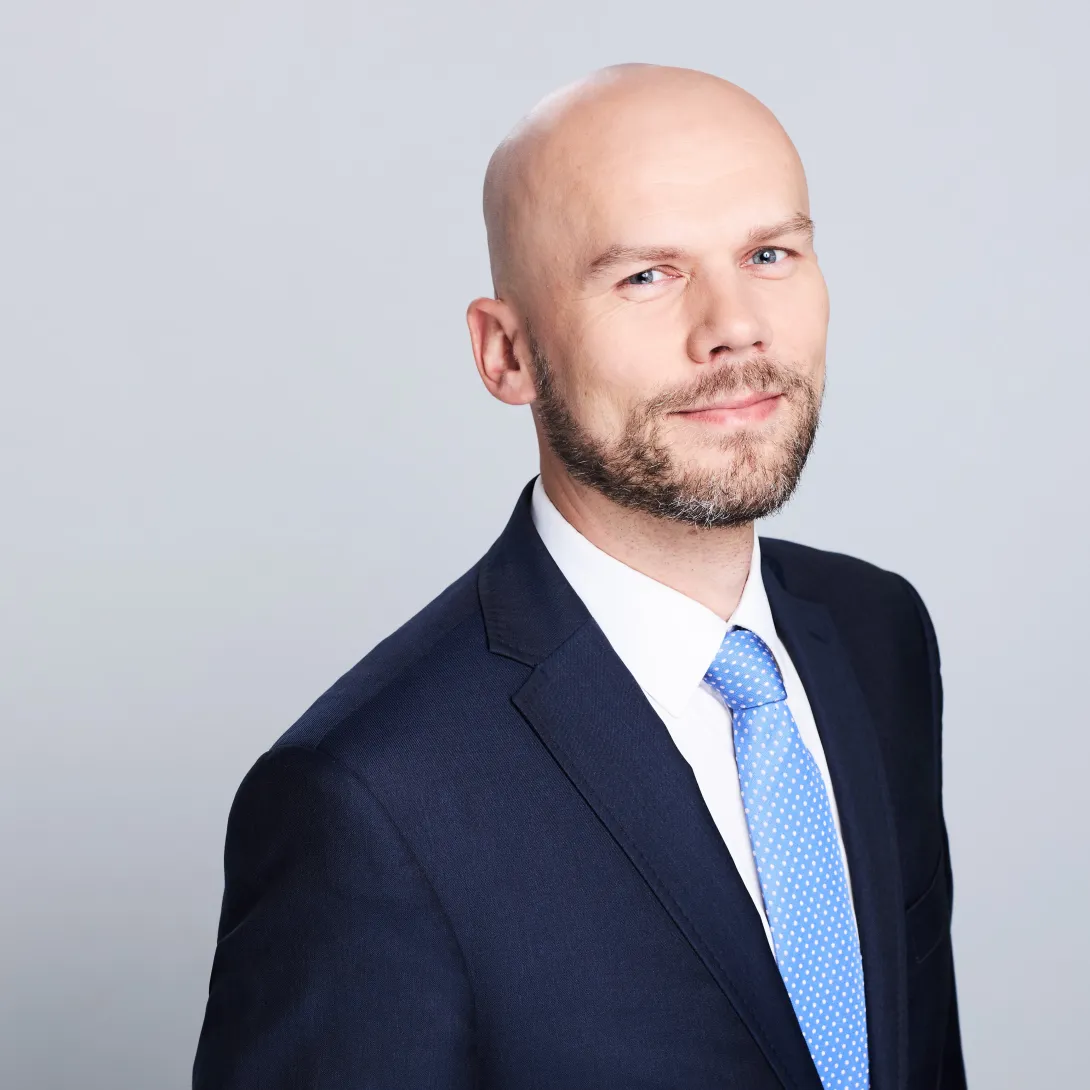 Stojący, łysy mężczyzna z brodą w granatowym garniturze na szarym tle