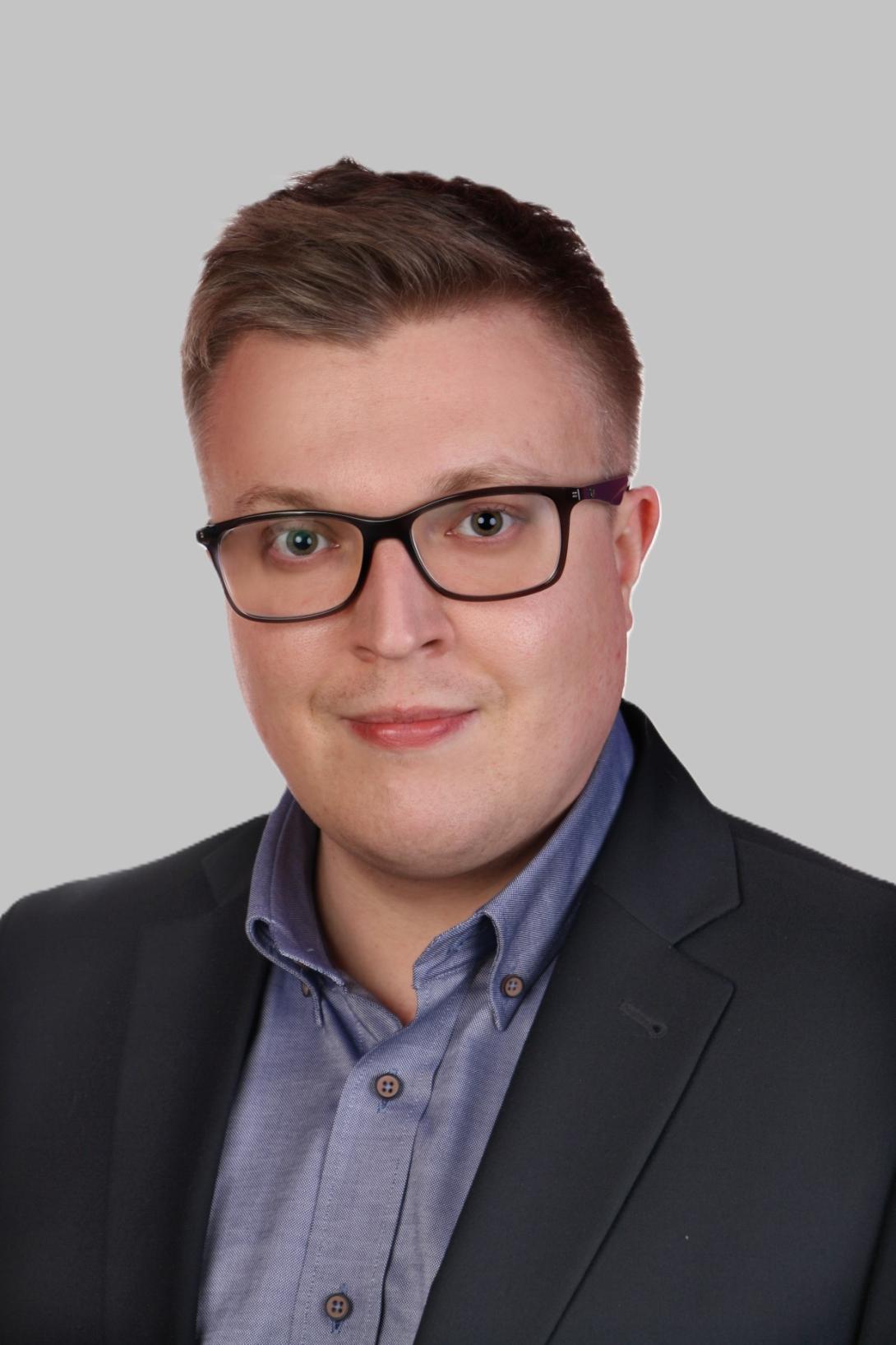 Uśmiechnięty mężczyzna w okularach, ubranym w ciemną marynarkę i błękitną koszulę, pozuje na jednolitym, jasnym tle.